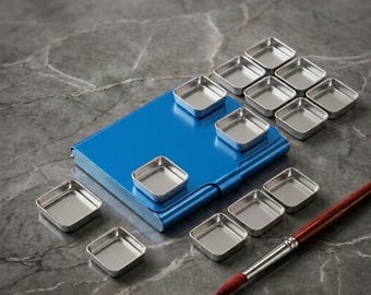Puede incluir: Una caja de metal azul brillante para acuarelas con tapa abatible, acompañada de doce godets cuadrados de metal plateado vacíos dispuestos alrededor de la caja y un pincel con mango de madera rojo oscuro y virola plateada.