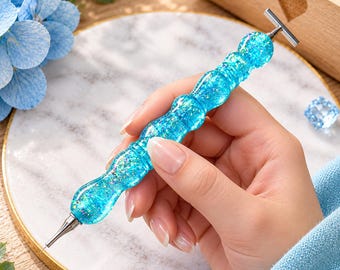 Puede incluir: Un bolígrafo para pintura de diamantes con un mango de resina azul brillante, translúcido y lleno de purpurina, con una forma ergonómica contorneada. La herramienta incluye una punta de metal plateado y un accesorio en forma de T.