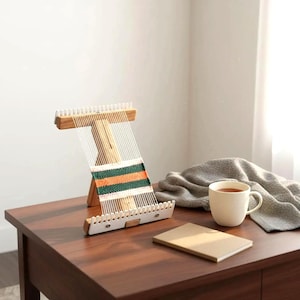 May include: A wooden weaving loom with white warp threads and a partially woven textile featuring horizontal stripes in green, orange, and white. The loom has a light wood frame with white notched plastic ends for securing the threads.
