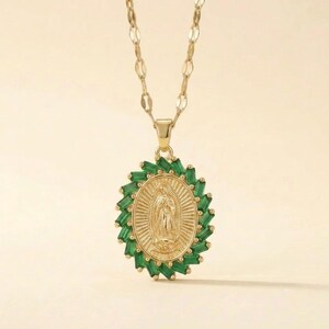 Puede incluir: Un collar dorado con un colgante ovalado que presenta una figura en relieve de la Virgen de Guadalupe. El colgante está rodeado por un halo de piedras verdes de corte baguette y cuelga de una delicada cadena de eslabones dorada.