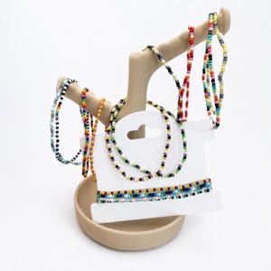 Könnte beinhalten: Eine Sammlung von mehrfarbigen Rocailles-Armbändern und Halsketten, die auf einem beigen, handförmigen Schmuckständer präsentiert werden. Der Schmuck besteht aus verschiedenen Mustern kleiner, leuchtender Glasperlen in Rot, Blau, Gelb, Grün und Weiß.