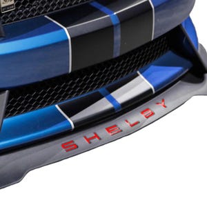May include: A blue car front bumper featuring a black honeycomb grille and a prominent black front splitter. The bumper displays a central black racing stripe with thin white and blue pinstripes. The word SHELBY is embossed in red lettering.