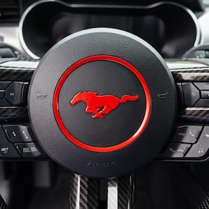 May include: A black leather steering wheel featuring a central circular emblem with a red running horse logo surrounded by a red ring. The steering wheel includes carbon fibre accents, integrated control buttons, and the word AIRBAG embossed below.