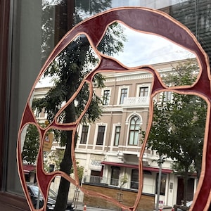 May include: A circular wall mirror featuring a segmented design with multiple reflective glass panels. The frame is constructed from dark red, opaque resin sections separated by thin, raised copper-colored metal lines in a web-like pattern.