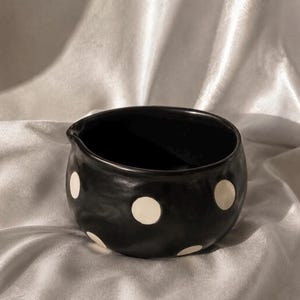 May include: A small, round, black ceramic bowl featuring a smooth, matte finish and a series of evenly spaced, circular white polka dots. The bowl has a distinct, subtle pouring spout integrated into its upper rim, creating a functional design.