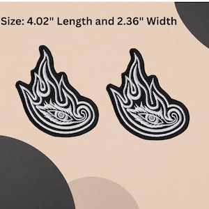 Puede incluir: Dos parches bordados en blanco y negro que presentan un ojo estilizado rodeado de patrones en forma de llama. El texto Size: 10.21 cm Length and 5.99 cm Width es visible. Cada parche tiene un fondo negro con hilo blanco detallando el ojo.