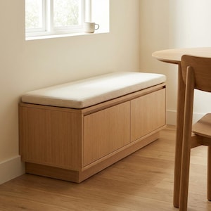 May include: A rectangular light oak storage bench featuring two cabinet doors and a thick, off-white rectangular cushion on top. The bench is positioned against a wall near a window, with a wooden chair leg visible in the foreground.