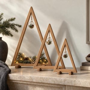 May include: Three triangular wooden frames of varying heights, each featuring a clear glass ornament hanging from the top apex. The ornaments contain small pinecones and greenery. The base of each frame is decorated with pine branches and fairy lights.