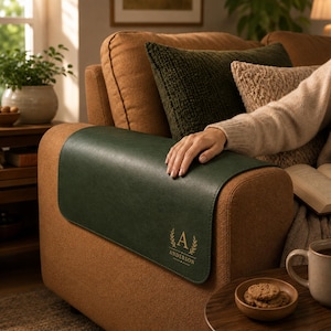May include: A dark green rectangular armrest cover made of a smooth material, featuring a gold-colored monogram with the letter A surrounded by a laurel wreath design and the text ANDERSON printed underneath, resting on a brown fabric sofa arm.