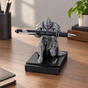 May include: A silver-colored knight figurine kneeling on a black rectangular base, holding a dark blue ballpoint pen with the text "Your Name" printed in white. The base features a white letter "J" and a series of rectangular indentations.