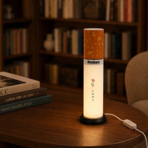 May include: A cylindrical table lamp designed to resemble a cigarette, featuring a textured orange filter top, a white body with the text Marlboro, and a small floral graphic above the name Emma printed vertically in black ink on the illuminated base.