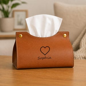 Puede incluir: Una funda rectangular para caja de pañuelos de color tostado con un icono de corazón negro centrado sobre el nombre Sophia. La funda tiene costuras visibles en los bordes y dos botones de presión metálicos dorados en las esquinas superiores.