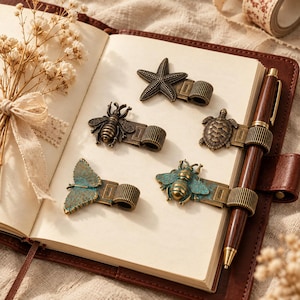 May include: A collection of five metal pen holders featuring antique-style designs including a starfish, a turtle, two bees, and a butterfly. The holders are attached to the pages of an open notebook, with one dark brown wooden pen secured in place.