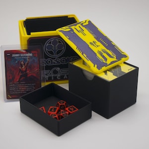 May include: A black and yellow gaming card storage box with a matching lid featuring a purple geometric pattern. A separate black tray contains several red and black multi-sided dice. A single trading card featuring Johnny Silverhand is included.
