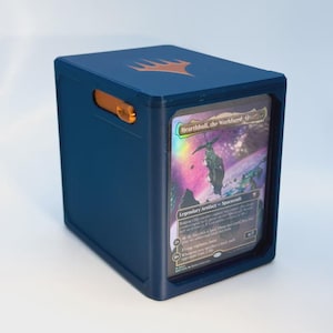 Op de afbeelding: Een donkerblauwe rechthoekige deckbox met een oranje Magic: The Gathering-logo op de bovenkant en een oranje zijsluiting. Een holografische Hearthhull, the Worldseed-verzamelkaart is zichtbaar in het doorzichtige venster aan de voorzijde.