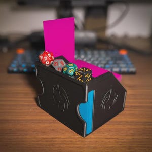 Puede incluir: Una caja de dados de juego de plástico negro con una tapa interior de color rosa brillante y un panel lateral azul. La caja presenta diseños circulares en relieve en el exterior y contiene cuatro dados poliédricos en rojo, gris, turquesa y negro.