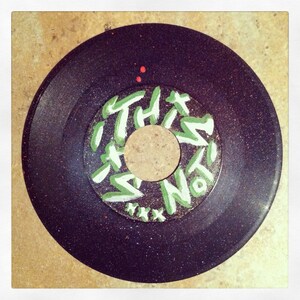 May include: A black vinyl record with a green and white label that says "This Is Not" with a white "xxx" underneath. The record has a speckled surface.