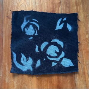 May include: A black fabric patch with a blue stencil design of stylized flowers. The flowers are outlined in black and have a light blue fill.