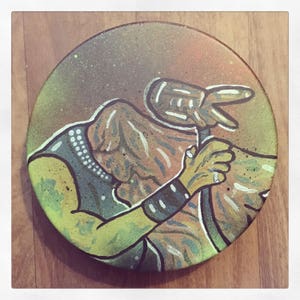 May include: A hand-painted ceramic plate featuring a colorful illustration of a person holding a microphone. The person is wearing a black and white striped shirt and has a green and yellow arm. The background is a light brown color.