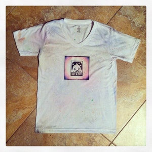 May include: White t-shirt with a black and white graphic of a panda bear with the text "NOT A TOY" printed on it. The shirt has a V-neck and is splattered with colorful paint.