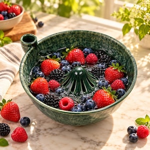 May include: A dark green ceramic berry colander bowl with a central raised drainage cone and a small side handle, filled with a mix of fresh red strawberries, dark blackberries, and small blue blueberries in water.