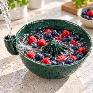 May include: A dark green speckled ceramic berry colander with a central cone and a side spout. Water pours into the spout, filling the bowl which contains fresh raspberries and blueberries. The bowl has a textured, glossy finish.