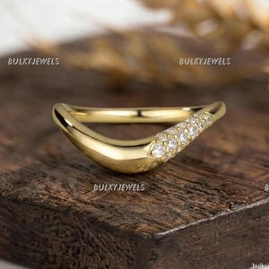 Può includere: Un anello in oro curvo caratterizzato da una fascia lucida che si restringe verso una sezione incastonata con piccole pietre chiare a taglio rotondo. L'anello ha una finitura liscia e brillante e un contorno a V, con il testo BULKYJEWELS visibile.