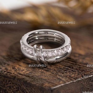 Puede incluir: Un anillo de tono plateado con una banda central incrustada con una hilera de piedras claras de corte redondo. El diseño incluye dos bucles plateados lisos y pulidos que se superponen a la banda principal. El texto BULKYJEWELS es visible.
