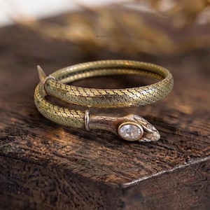 May include: A wrap-around snake ring crafted from textured gold-tone metal, featuring a detailed scale pattern along the band. The head of the snake is accented with a single, clear, oval-shaped gemstone set into the top, creating a coiled design.