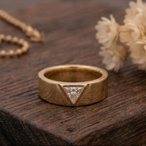 Pode incluir: Um anel de banda em ouro com uma única pedra preciosa transparente em forma de triângulo, engastada ao nível do centro da superfície metálica. O anel tem um acabamento liso e polido e um perfil largo e plano com um design geométrico.