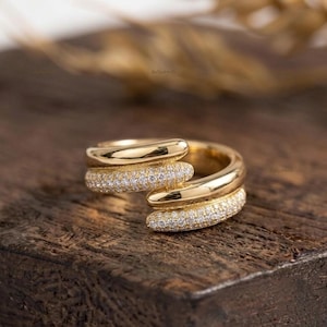 Puede incluir: Un anillo bypass de oro con dos bandas superpuestas. Una banda es de oro pulido liso, mientras que la otra está incrustada con pequeñas piedras claras en pavé. El diseño crea una apariencia abierta y superpuesta con un acabado metálico brillante.