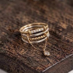 Op de afbeelding: Een ring met meerdere banden in een combinatie van goud- en zilverkleurig metaal. De bovenste band is versierd met vijf kleine heldere steentjes, terwijl een fijn gouden kettinkje twee banden verbindt en een peervormige bedel met steen draagt.