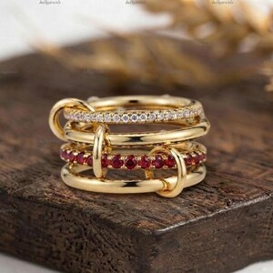 Könnte beinhalten: Ein mehrreihiger Goldring bestehend aus vier miteinander verbundenen Ringen. Ein Ring ist mit klaren weißen Steinen besetzt, während ein anderer eine Reihe leuchtend roter Steine aufweist. Die Ringe sind durch kleine goldene Schlaufen verbunden.