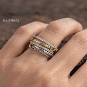Op de afbeelding: Een ring met meerdere banden bestaande uit een combinatie van gepolijste goudkleurige en zilverkleurige metalen banden, waarvan twee banden zijn ingelegd met kleine heldere steentjes. Het ontwerp bestaat uit in elkaar grijpende elementen.