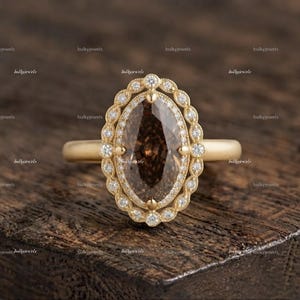 Puede incluir: Un anillo de oro con una gran piedra preciosa marrón central de corte marquesa, rodeada por un halo festoneado de pequeños diamantes blancos redondos y claros. La banda es de oro liso y pulido, creando un contraste con la piedra central oscura y facetada.