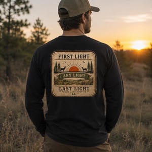 May include: A black long-sleeve shirt featuring a rectangular graphic on the back. The design displays a sunrise over a forest with two deer, the text FIRST LIGHT, ANY LIGHT, LAST LIGHT, and PUBLIC LAND HUNTING, framed by a stitched border.