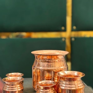 Può includere: Un set di quattro recipienti in rame lucido composto da una grande pentola centrale e tre pentole più piccole abbinate. Ogni recipiente presenta un bordo largo e svasato, un corpo arrotondato e una superficie metallica liscia e riflettente di colore bruno-rossastro.