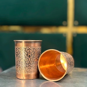 Puede incluir: Dos vasos cilíndricos de cobre con un intrincado patrón floral oscuro grabado en la superficie exterior. Un vaso está en posición vertical, mientras que el otro está inclinado de lado, revelando el acabado interior metálico liso y pulido.