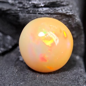Puede incluir: Una piedra preciosa de ópalo tallada en cabujón redondo con un color de cuerpo naranja pálido y translúcido. La piedra muestra destellos internos de color iridiscente, incluyendo reflejos de luz amarilla, naranja y verde brillante en su superficie.