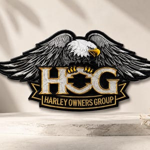 May include: A decorative wall sign featuring a bald eagle with spread wings perched above the letters HOG in a metallic gold and silver finish. Below the letters, a black banner displays the text HARLEY OWNERS GROUP in gold lettering.