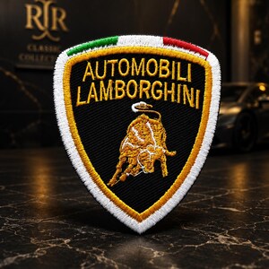 May include: A shield-shaped embroidered patch featuring the Automobili Lamborghini logo. The design includes a black background with a gold bull emblem, yellow border, and a top stripe in green, white, and red. The text Automobili Lamborghini is in gold.