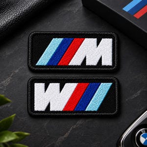 May include: Two rectangular embroidered patches featuring the M logo. Each patch has a black background with a white letter M followed by three diagonal stripes in light blue, dark blue, and red. The embroidery is dense with a stitched border.