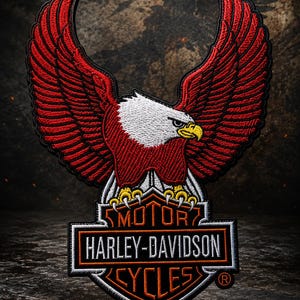 May include: An embroidered patch featuring a bald eagle with large red wings spread wide, perched atop a black and orange Harley-Davidson Motor Cycles logo. The eagle has a white head and a yellow beak, with the brand name clearly stitched in white text.