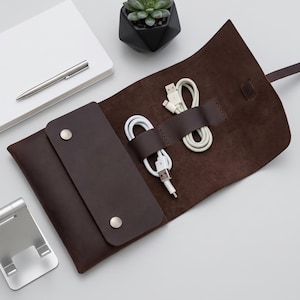 May include: A dark brown leather roll-up organizer featuring a flap secured with two silver-toned metal snaps. The interior includes two leather loops holding white charging cables with USB connectors, providing a compact storage solution.