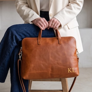 Könnte beinhalten: Eine hellbraune Laptoptasche aus Leder mit zwei Tragegriffen und einem abnehmbaren Schultergurt. Die rechteckige Tasche hat eine glatte Oberfläche und ist in der unteren rechten Ecke mit den goldenen Initialen KL geprägt.