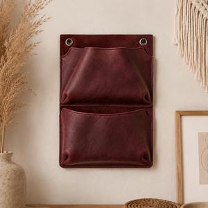 May include: A deep burgundy leather wall organiser featuring two stacked open pockets. The rectangular piece is secured at the top with two metallic grommets, providing a structured storage solution with a smooth, matte finish and visible stitching.