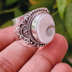 Pode incluir: Um anel de prata com uma grande concha de olho de shiva branca de formato oval com um padrão espiral natural. A banda de metal é decorada com intrincados detalhes de contas de prata e texturas de corda ao redor do engaste central.