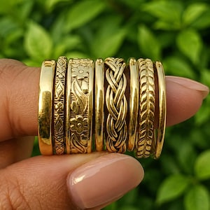 May include: A set of seven gold-toned rings featuring various designs including a smooth band, a geometric pattern, a floral motif, a plain thin band, a braided texture, a leaf-like pattern, and a simple thin band, all displayed together.