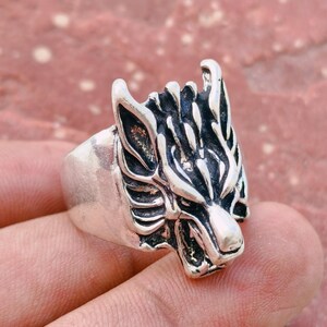 Puede incluir: Un anillo de metal en tono plateado con un diseño detallado y esculpido de una cabeza de lobo. La superficie del anillo tiene un acabado pulido en las áreas elevadas y una textura oscura y hundida para resaltar los rasgos faciales del lobo.