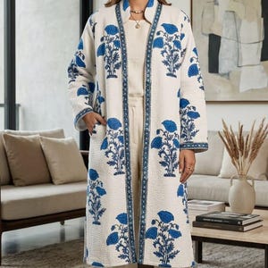 May include: A long, cream-colored quilted coat featuring a vibrant blue floral print. The coat has an open front, long sleeves, and side pockets. It is worn over a matching cream-colored top and straight-leg trousers, paired with brown sandals.